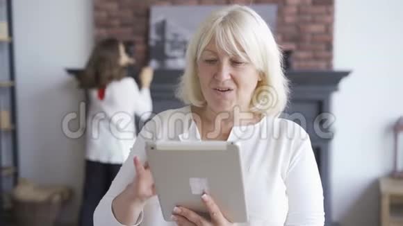 一幅成熟的女人坐在沙发上看着平板电脑上的照片而她的女儿则在打扫房间妇女视频的预览图