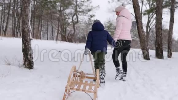 穿着温暖衣服的可爱男孩和他的母亲在下雪的冬日拉着木制的老式雪橇散步一个小男孩滑了进去视频的预览图