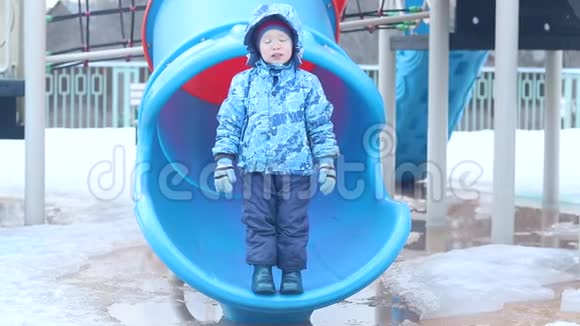 在操场上穿着橡胶靴的小男孩从一座小山跳到一个水坑里春天冰在融化水在周围视频的预览图
