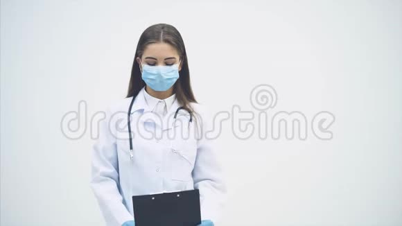 年轻的女实习生站在医疗制服和面具拿着剪贴板检查数据摇头翻转视频的预览图