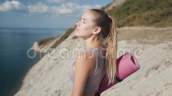 女人拿着瑜伽垫从悬崖上看海深呼吸慢动作视频的预览图
