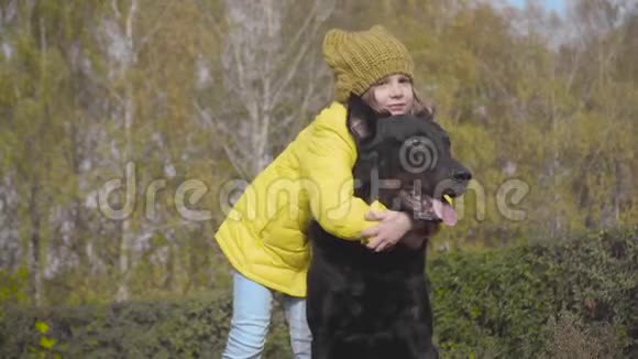 穿着芥末帽和黄色外套的白人女孩微笑着拥抱秋天公园里的狗孩子们和她一起度过周末视频的预览图