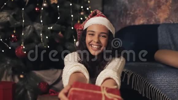 带上心满意足的年轻女孩戴着桑塔帽和白色套头衫伸出红色礼品盒微笑着拍照视频的预览图