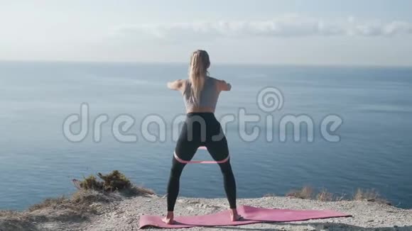 运动的女人在海洋附近用橡皮筋做蹲姿动作缓慢视频的预览图