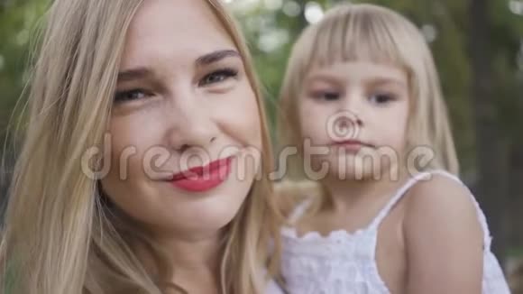 一幅年轻的金发女人抱着她可爱的女儿穿着白色连衣裙抱着她的女儿看着镜头户外休闲视频的预览图