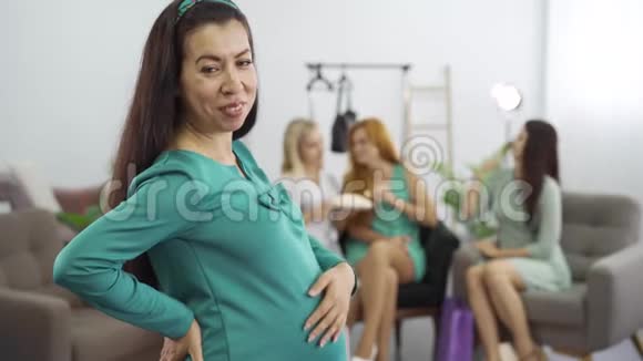 微笑着穿着蓝绿色连衣裙的孕妇抚摸着她的腹部准妈妈和她的三个朋友在网上聊天视频的预览图