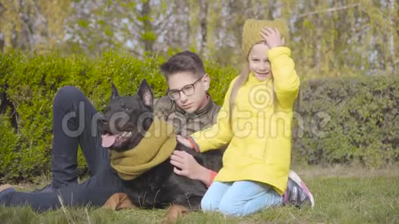 戴眼镜的白种人少年和穿黄色外套的妹妹坐在绿草上戴着漂亮的杜宾视频的预览图