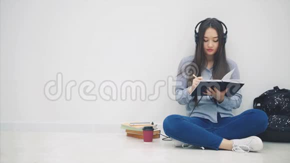 有趣的年轻亚洲女孩坐在地板上戴着耳机听着音乐停止写作自拍视频的预览图