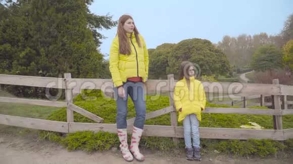 母亲和女儿穿着类似的黄色外套和牛仔裤站在秋天公园的栅栏旁边望着远方视频的预览图