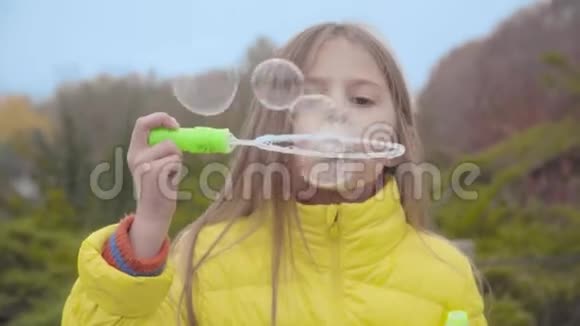 可爱的白人女孩长发和棕色的眼睛吹肥皂泡在户外在秋天公园休息的漂亮孩子视频的预览图