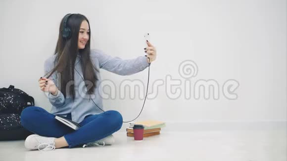 有趣的年轻亚洲女孩坐在地板上戴着耳机听着音乐自拍摆姿势挥手问好视频的预览图