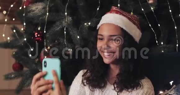 圣诞老人帽子里的年轻女孩坐在装饰好的圣诞树下用她的智能手机进行视频聊天视频的预览图