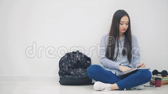 充满灵感的年轻亚洲女孩坐在地板上完成写作拿起电话和耳机正在听音乐视频的预览图