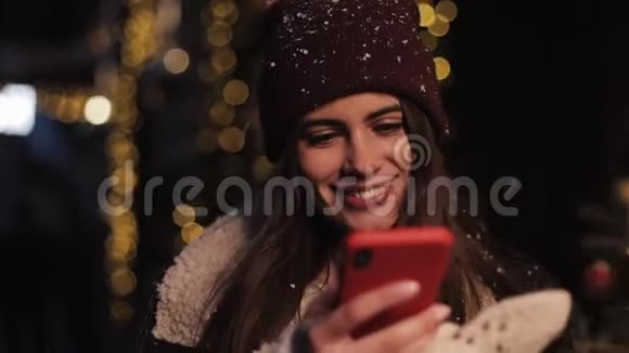 在社交网络上戴着冬帽的微笑迷人的女孩用智能手机看着屏幕视频的预览图