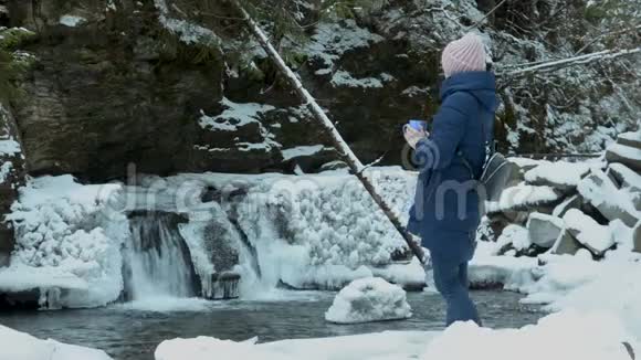 穿着蓝色外套的女孩站在冰雪覆盖的针叶林中脸上挂着一个杯子背景是冰冷的瀑布和岩石冬季视频的预览图