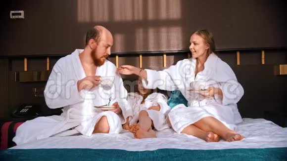 一个穿着白色浴袍的成功家庭坐在酒店的床上喝茶视频的预览图
