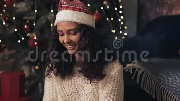 圣诞老人帽中年轻漂亮的穆拉托女孩坐在装饰的圣诞树下准备礼物包装视频的预览图