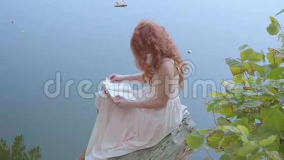 侧面看一个年轻的白种人女人留着长长的红色头发在蓝湖的背景下阅读这本书可爱的女孩视频的预览图