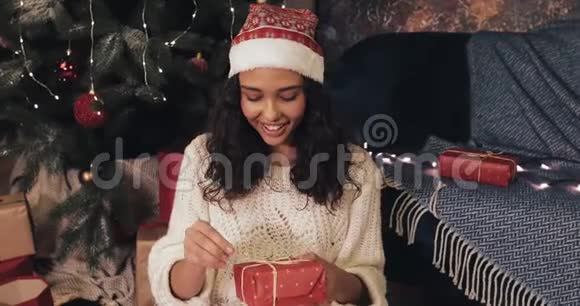 美丽的微笑穆拉托女孩坐在圣诞树下舒适的家背景同时解开红色礼物盒视频的预览图