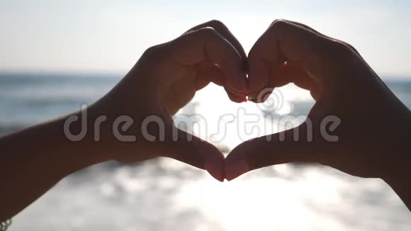 年轻女孩在日出时用双手在海洋背景上做心形海滩上的暑假假期概念视频的预览图
