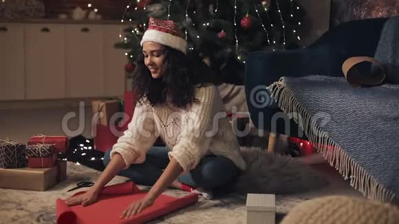 年轻漂亮的桑塔斯帽木乃托女孩坐在装饰过的圣诞树下展开纸卷把视频的预览图