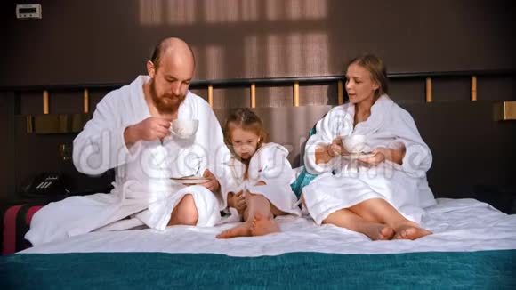 一个成功的幸福家庭穿着白色浴袍坐在酒店的床上喝着茶视频的预览图