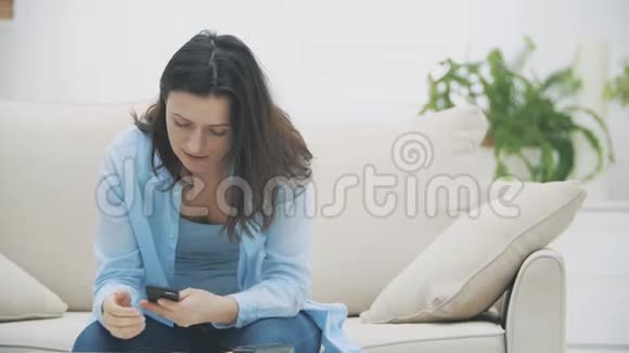 令人钦佩的女人专注于使用她的智能手机而坐在沙发上在家里慢动作复制空间4K视频的预览图