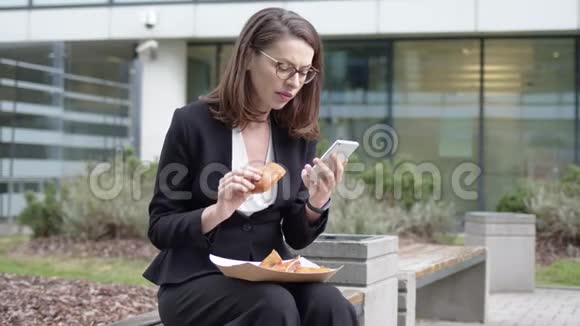 女商人在院子里浏览智能手机和吃三明治视频的预览图