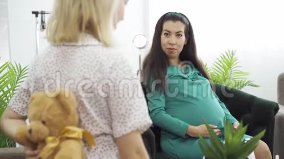 穿着浅色礼服的女孩送给她的朋友一份礼物送给她未出生的孩子年轻的黑发孕妇蓝绿色视频的预览图