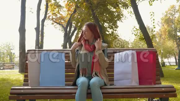 一个长着狐臭头发的女孩坐在公园的长凳上在去商店后用纸袋审视自己的购物视频的预览图