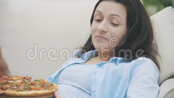 慢点一个年轻的女人坐在家里的沙发上一边吃披萨一边看电视4K视频的预览图