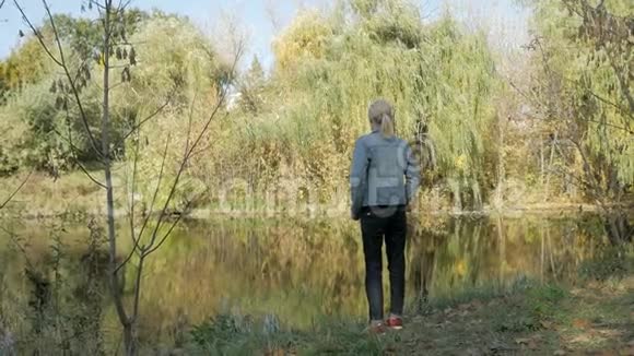 一个女人站在河岸上在晴朗的秋天看着远处从后面看到的视频的预览图