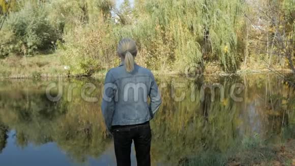 一个女人站在河岸上在晴朗的秋天看着远处从后面看到的视频的预览图