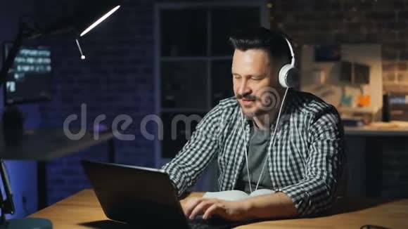 一个微笑的家伙晚上在办公室用笔记本电脑用耳机听音乐视频的预览图