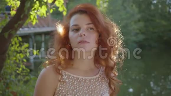 美丽苗条的白种人的肖像红色的头发穿着米色的连衣裙站在花园里看着远处连接视频的预览图