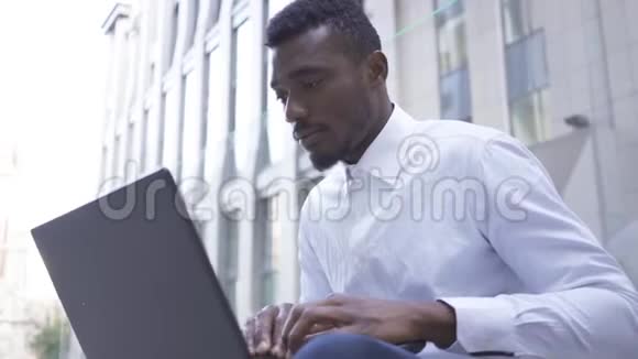 英俊的非洲裔美国人的肖像穿着白色衬衫坐在城市街道上的笔记本电脑上打字自信的男性视频的预览图