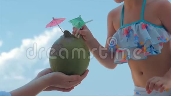 美丽的白种人小女孩穿着泳衣在海滩上蓝天喝着一管椰子饮料视频的预览图