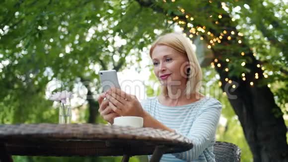 漂亮的成熟金发女郎使用智能手机在露天咖啡馆享受夏日视频的预览图