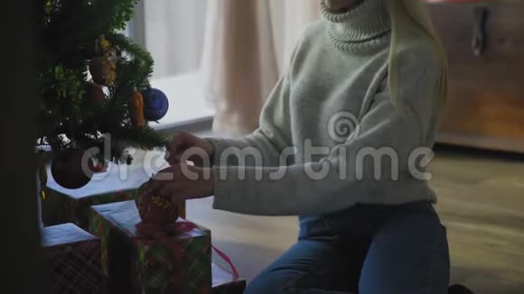 穿着毛衣和蓝色牛仔裤的年轻女人坐在地板上在家里用球装饰圣诞树视频的预览图