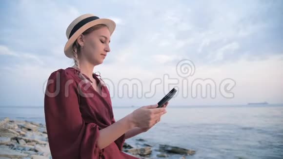 一个漂亮的女孩穿着一件夏天的红色连衣裙和一顶草帽晚上坐在海边的石头上视频的预览图