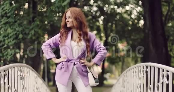 年轻的商务女性一头鲜红的头发正在夏季公园的一座步行桥上摆着相机视频的预览图