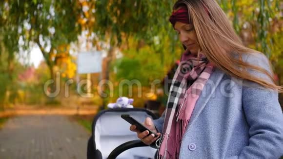 年轻快乐的母亲站在公园里秋天带着婴儿车在她的电话里拨了一条信息中弹视频的预览图