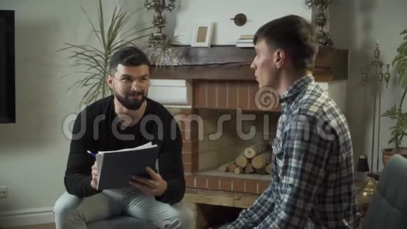 男性高加索心理学家戴着眼镜拿着笔记和他悲伤的病人交谈年轻男子视频的预览图