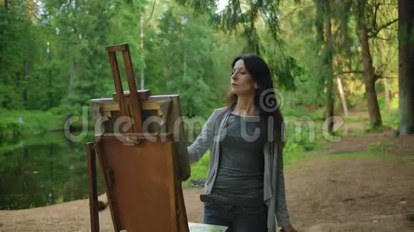 一位穿着衬衫和牛仔裤的漂亮女艺术家在池塘附近的公园里画一幅风景画视频的预览图