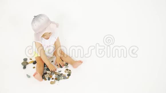 戴着斗笠的金发小女孩在白色背景上玩海鹅卵石和贝壳视频的预览图