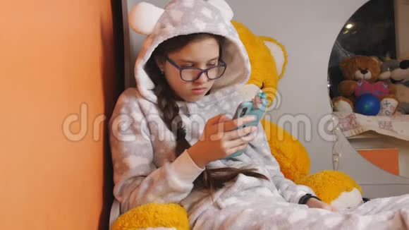 少女穿着温暖的睡衣躺在床上用智能手机和吃橘子视频的预览图