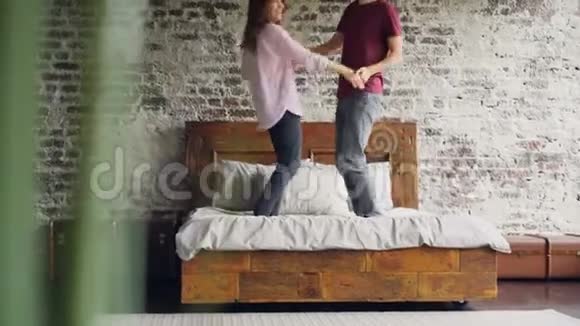 年轻的丈夫和妻子穿着休闲服装在床上跳跃和跳舞在漂亮的阁楼风格中欢笑和快乐视频的预览图