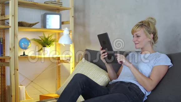 一位年轻的金发休闲女士正在自己的平板电脑上的客厅里度过一些空闲时间视频的预览图