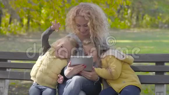 年轻的白种人带着一头金色卷发坐在秋天公园的长凳上和两个漂亮的小女孩在一起视频的预览图