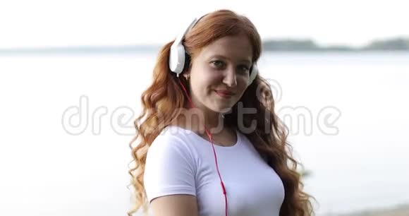把脸贴紧带着白色耳机的红发雀斑女孩正在湖畔听歌跳舞视频的预览图
